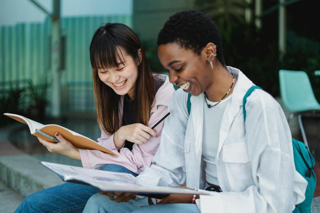 Two students in their late teens or early-mid twenties sit side by side smiling and holding notebooks in their hands.
